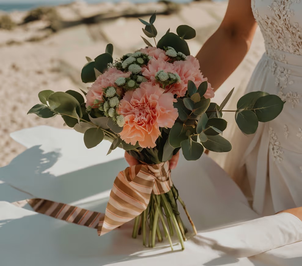 Ramo de novia con claveles rosados y eucalipto atado con cinta a rayas, sobre la playa